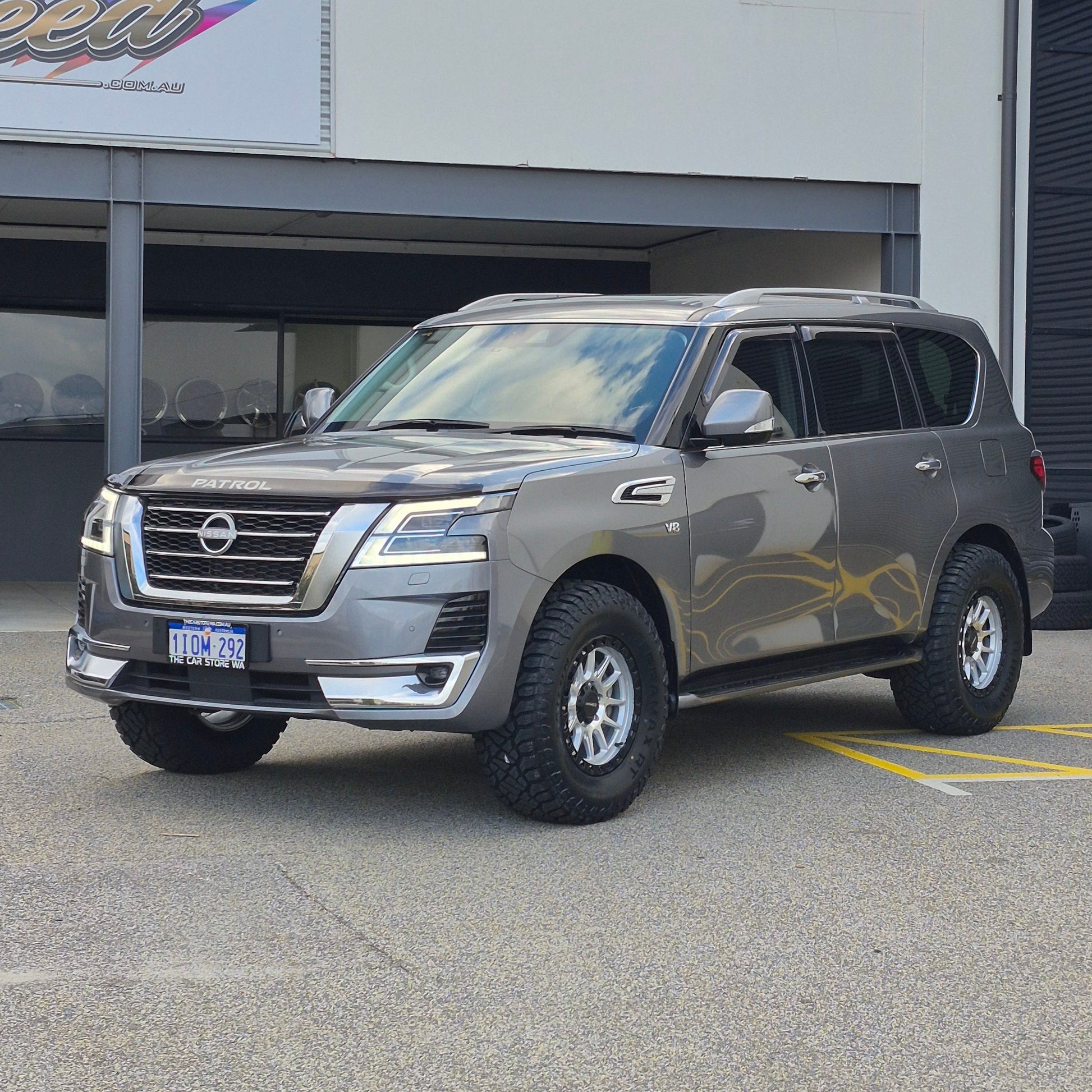 A dark gray Nissan y62 Patrol parked diagonally in front of a white building with large glass windows. DSpeed d-speed xt-1 xt1 18x9 4x4 wheels with 35in 35x12.5r18 RT tyres offroad 4x4 off road