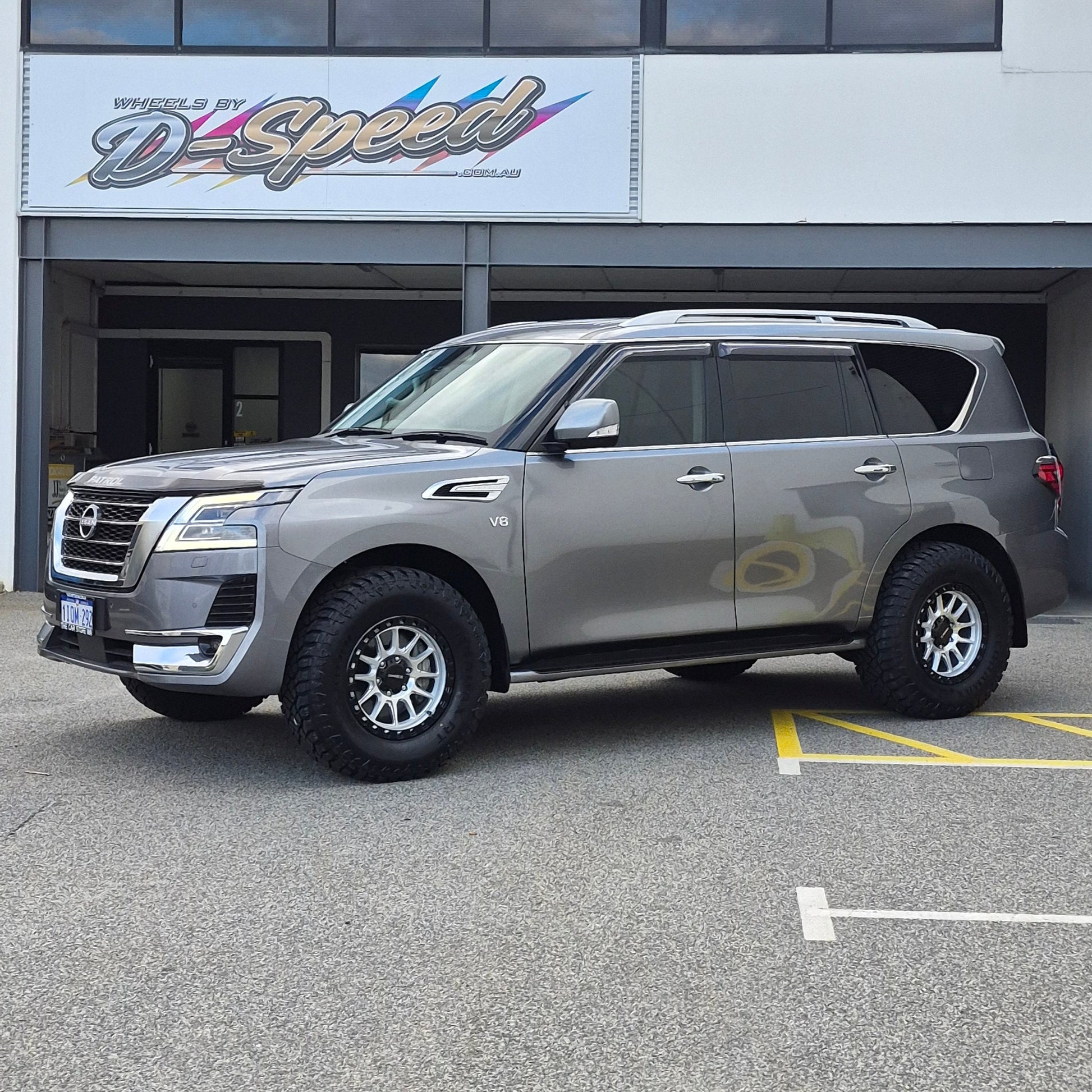 A dark gray Nissan y62 Patrol parked diagonally in front of a white building with large glass windows. DSpeed d-speed xt-1 xt1 18x9 4x4 wheels with 35in 35x12.5r18 RT tyres offroad 4x4 off road