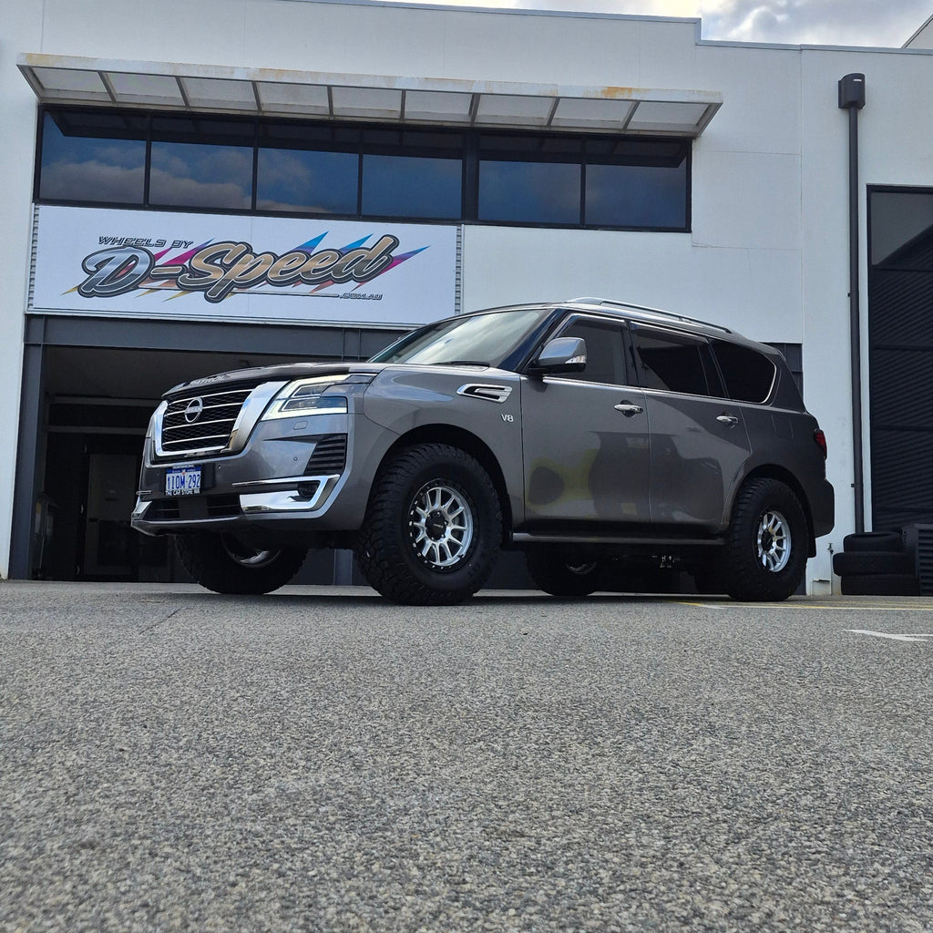 A dark gray Nissan y62 Patrol parked diagonally in front of a white building with large glass windows. DSpeed d-speed xt-1 xt1 18x9 4x4 wheels with 35in 35x12.5r18 RT tyres offroad 4x4 off road