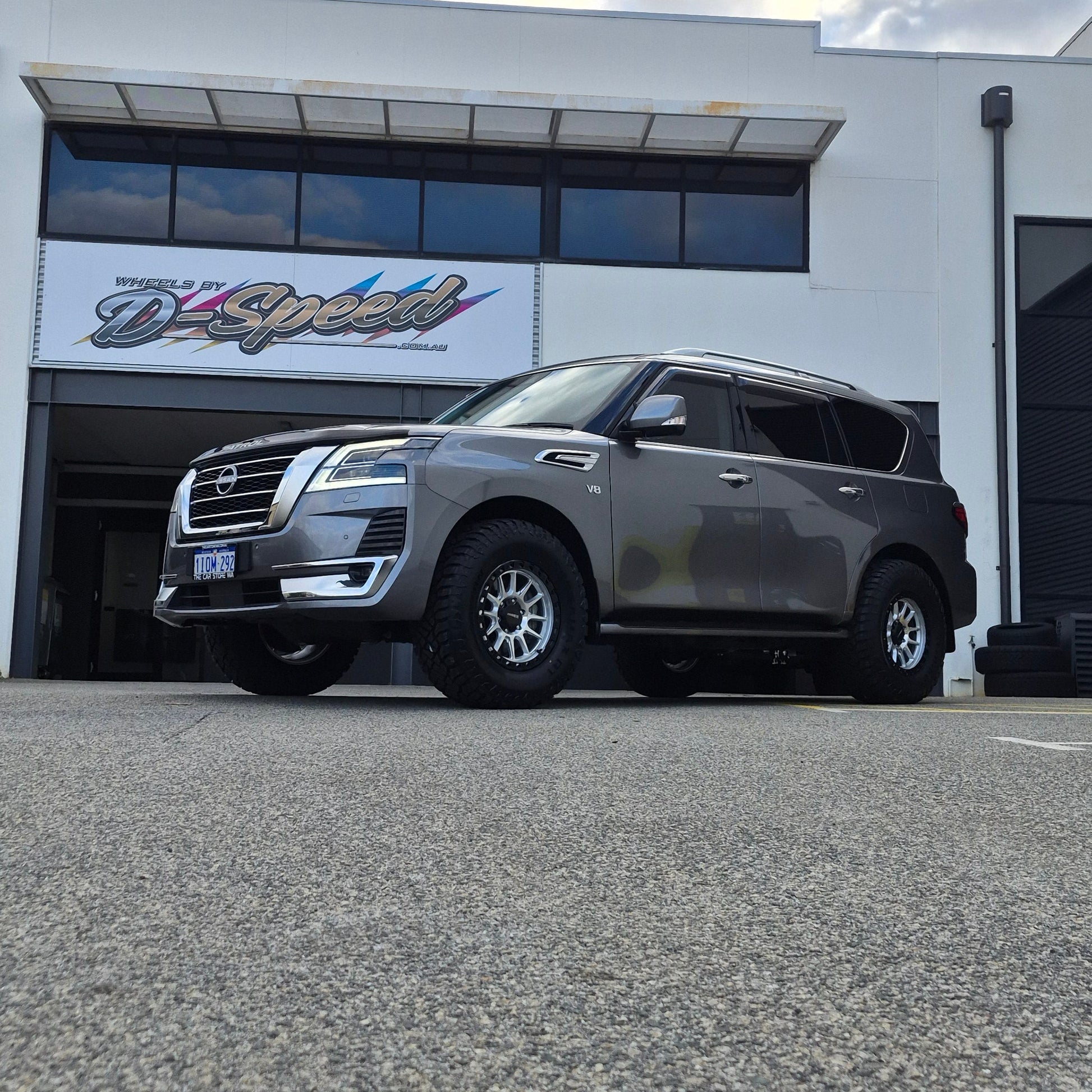 A dark gray Nissan y62 Patrol parked diagonally in front of a white building with large glass windows. DSpeed d-speed xt-1 xt1 18x9 4x4 wheels with 35in 35x12.5r18 RT tyres offroad 4x4 off road