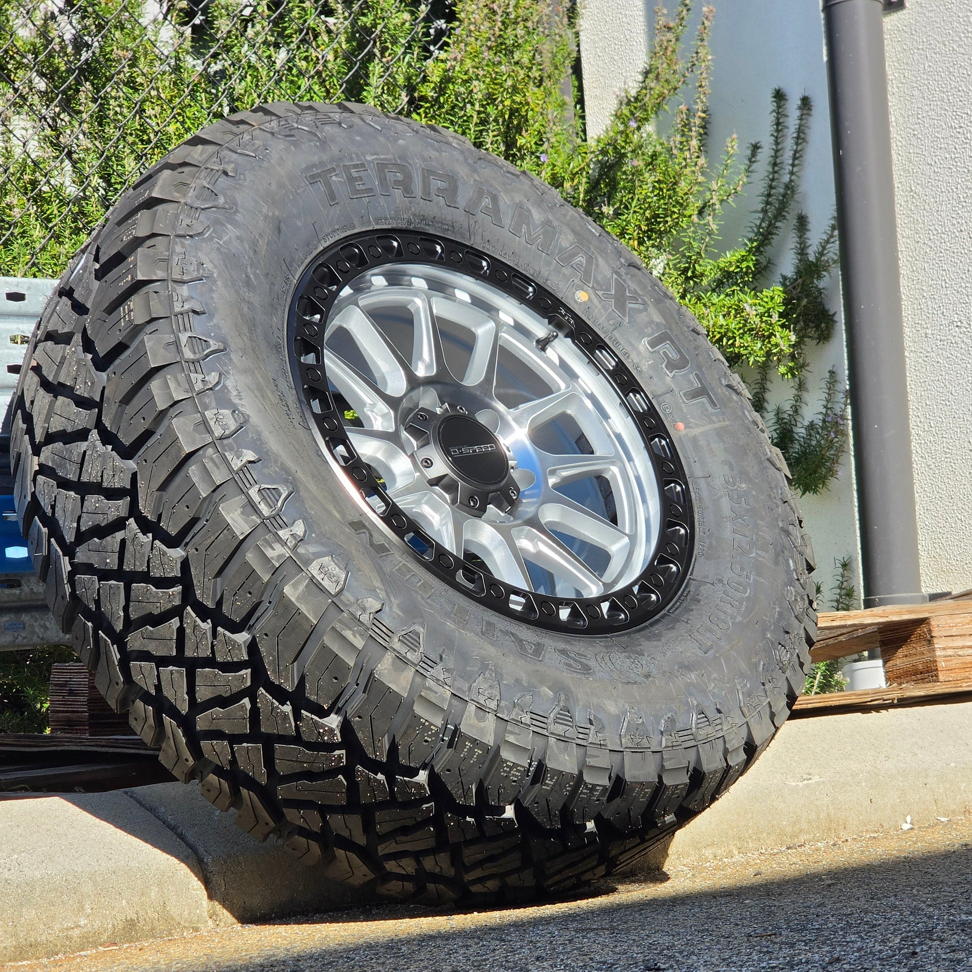 A black off-road tire with visible 'SAILUN terramax RT' lettering, mounted on a multi-spoke silver and black wheel, photographed outdoors against a chain-link fence and blue sky. Dspeed xt-1 xt1 wheel silver with black ring 17x9 17x8.5 18x9 4x4 offroad wheels rims