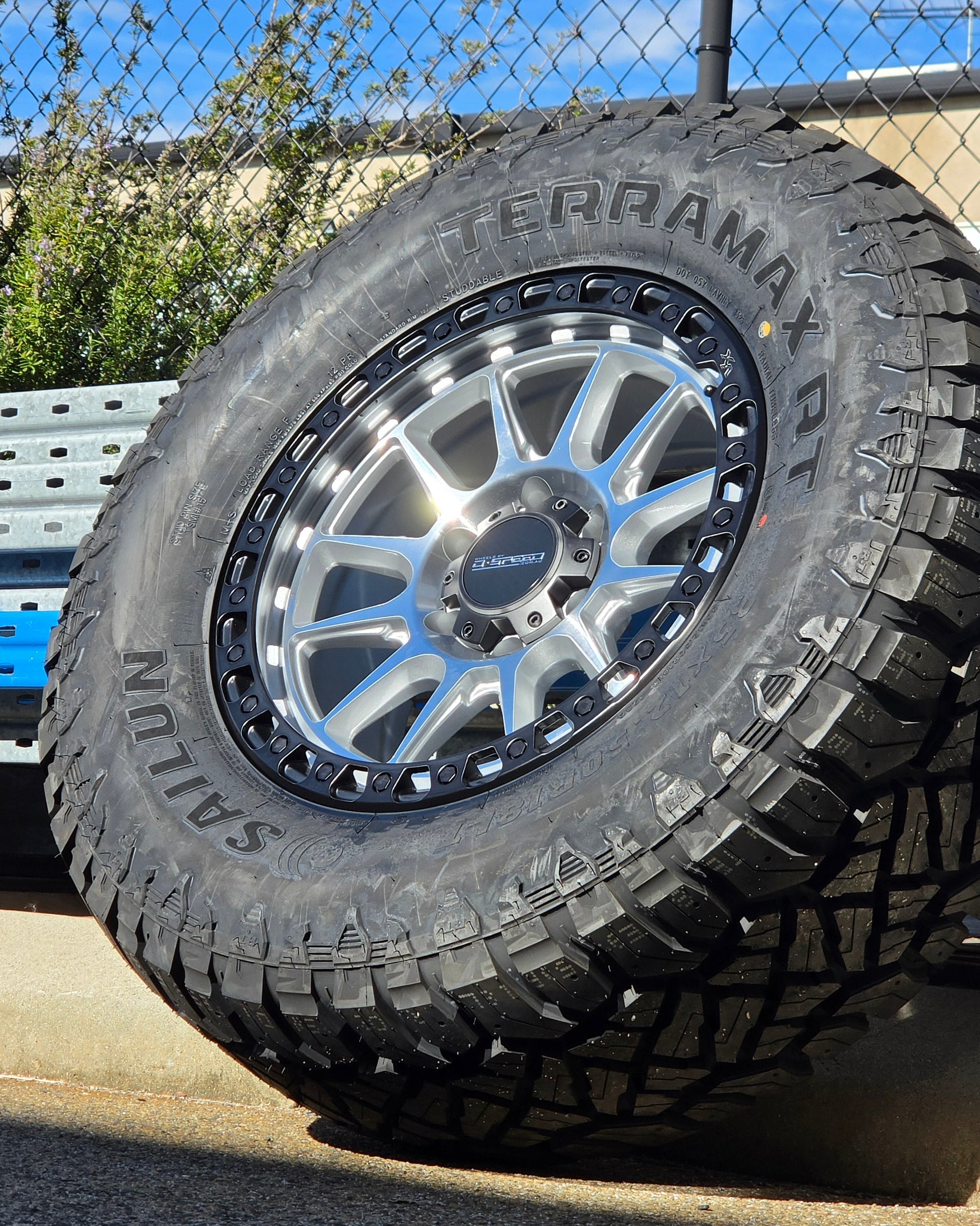 A black off-road tire with visible 'SAILUN terramax RT' lettering, mounted on a multi-spoke silver and black wheel, photographed outdoors against a chain-link fence and blue sky. Dspeed xt-1 xt1 wheel silver with black ring 17x9 17x8.5 18x9 4x4 offroad wheels rims