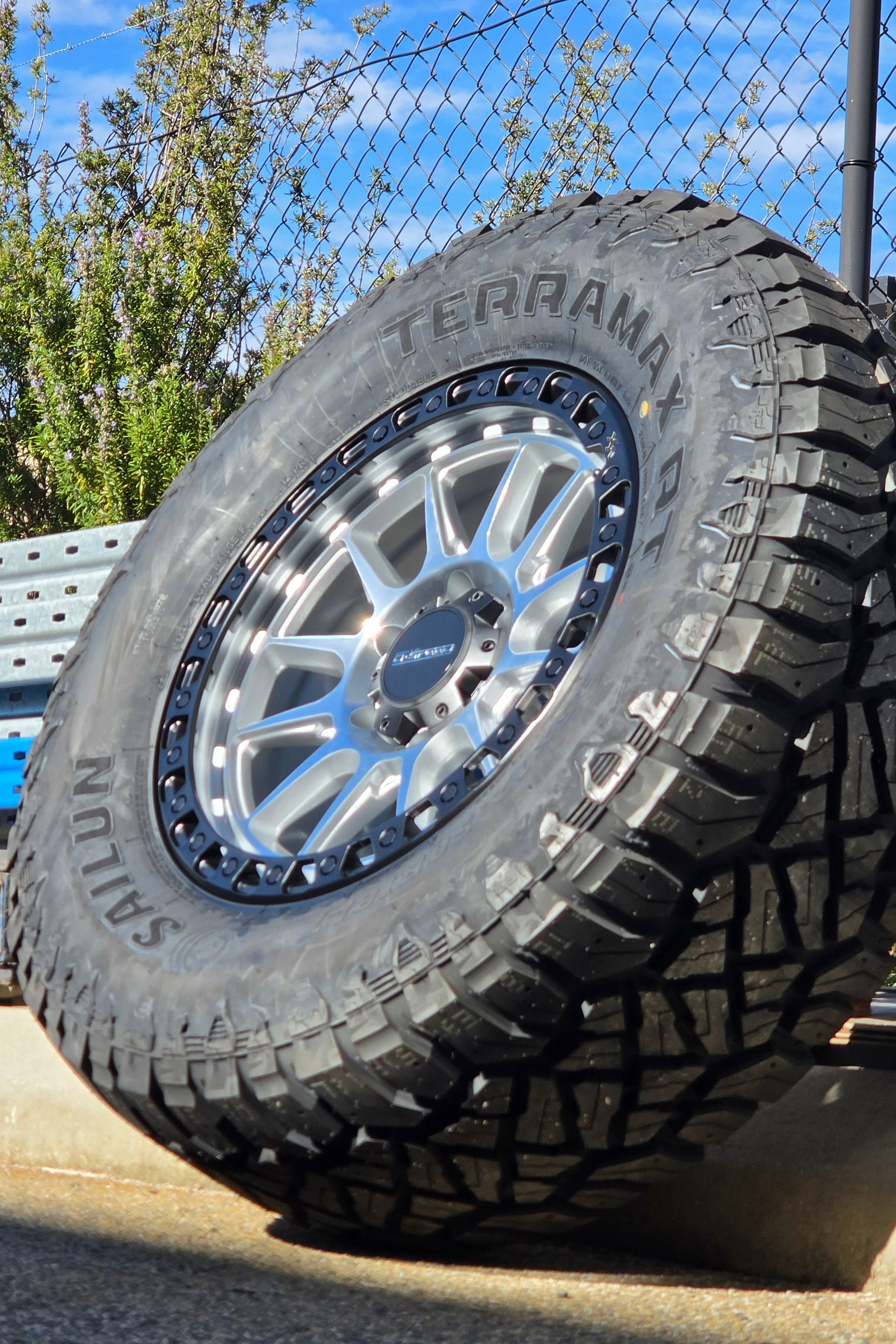 A black off-road tire with visible 'SAILUN terramax RT' lettering, mounted on a multi-spoke silver and black wheel, photographed outdoors against a chain-link fence and blue sky. Dspeed xt-1 xt1 wheel silver with black ring 17x9 17x8.5 18x9 4x4 offroad wheels rims