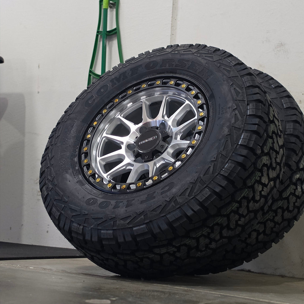 A large off-road tire mounted on a silver multi-spoke wheel, leaning against a gray wall in an indoor space. DSPEED D-SPEED XT-1 MACHINED SILVER WITH BLACK RING GOLD BOLTS 4X4 OFF ROAD OFFROAD WHEELS RIMS