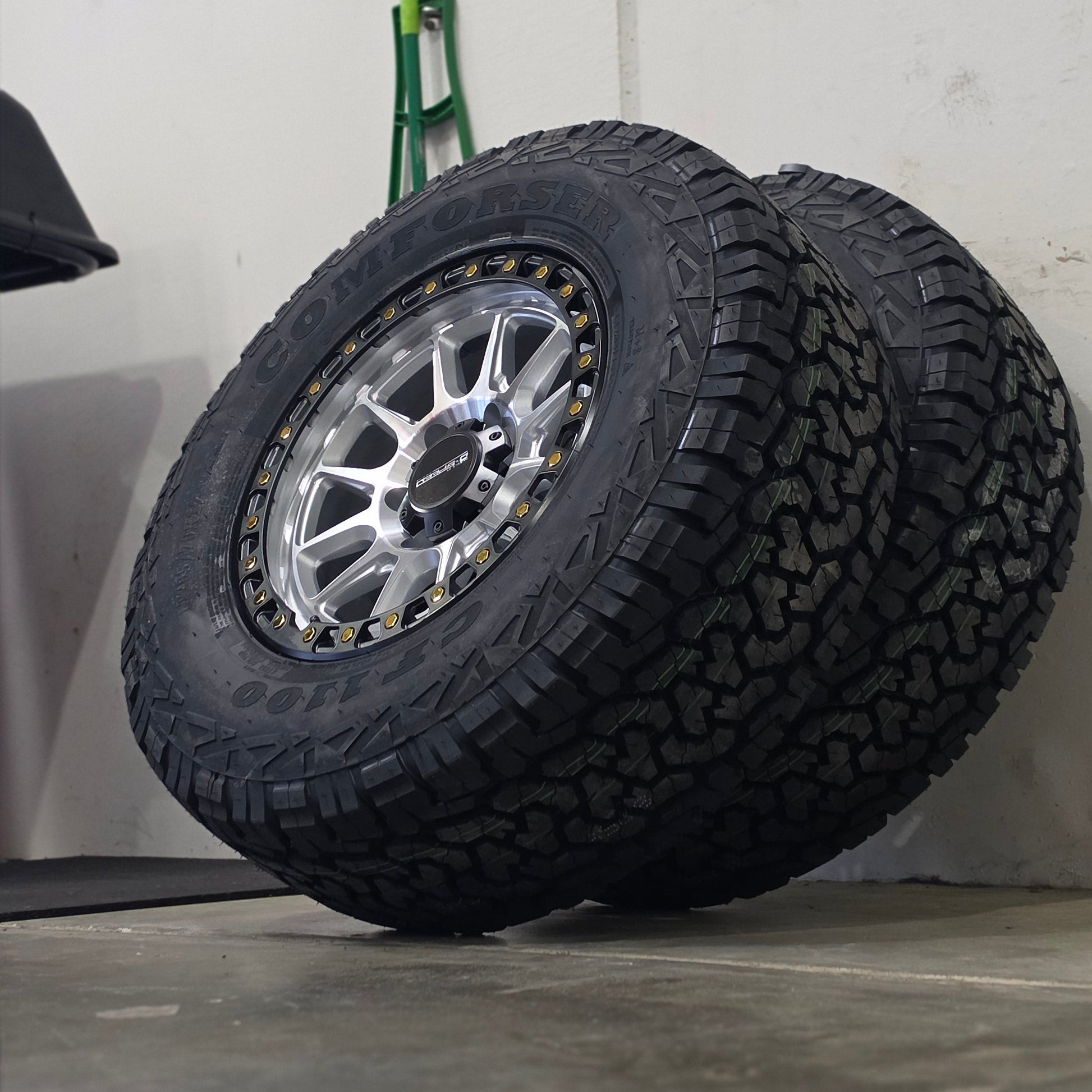 A large off-road tire mounted on a silver multi-spoke wheel, leaning against a gray wall in an indoor space. DSPEED D-SPEED XT-1 MACHINED SILVER WITH BLACK RING GOLD BOLTS 4X4 OFF ROAD OFFROAD WHEELS RIMS