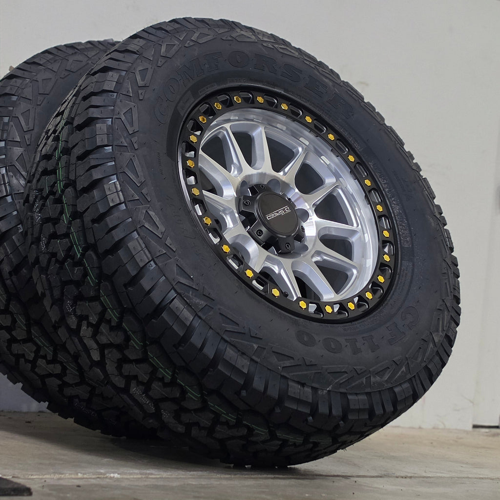 A large off-road tire mounted on a silver multi-spoke wheel, leaning against a gray wall in an indoor space. DSPEED D-SPEED XT-1 MACHINED SILVER WITH BLACK RING GOLD BOLTS 4X4 OFF ROAD OFFROAD WHEELS RIMS