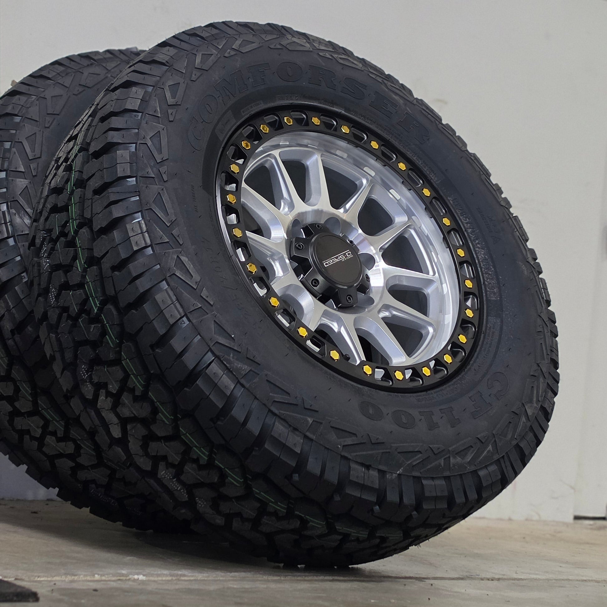 A large off-road tire mounted on a silver multi-spoke wheel, leaning against a gray wall in an indoor space. DSPEED D-SPEED XT-1 MACHINED SILVER WITH BLACK RING GOLD BOLTS 4X4 OFF ROAD OFFROAD WHEELS RIMS