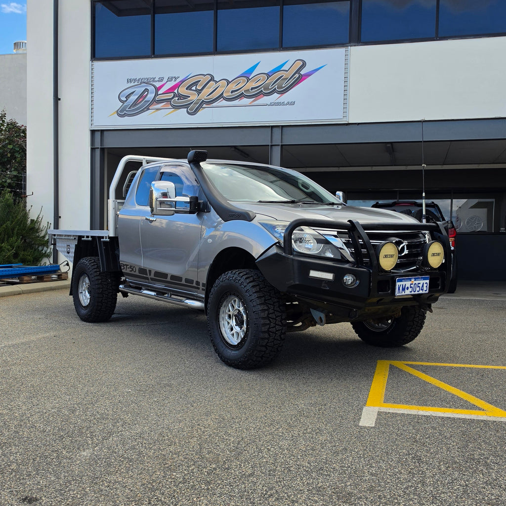 A dark gray off-road BT-50 with DSpeed MH-1 D-Speed MH1 4x4 off road offroad wheels 17x8.5 machined silver black Ring custom flatbed and front bumper guard parked in a paved lot in front of a beige commercial building under a blue sky.
