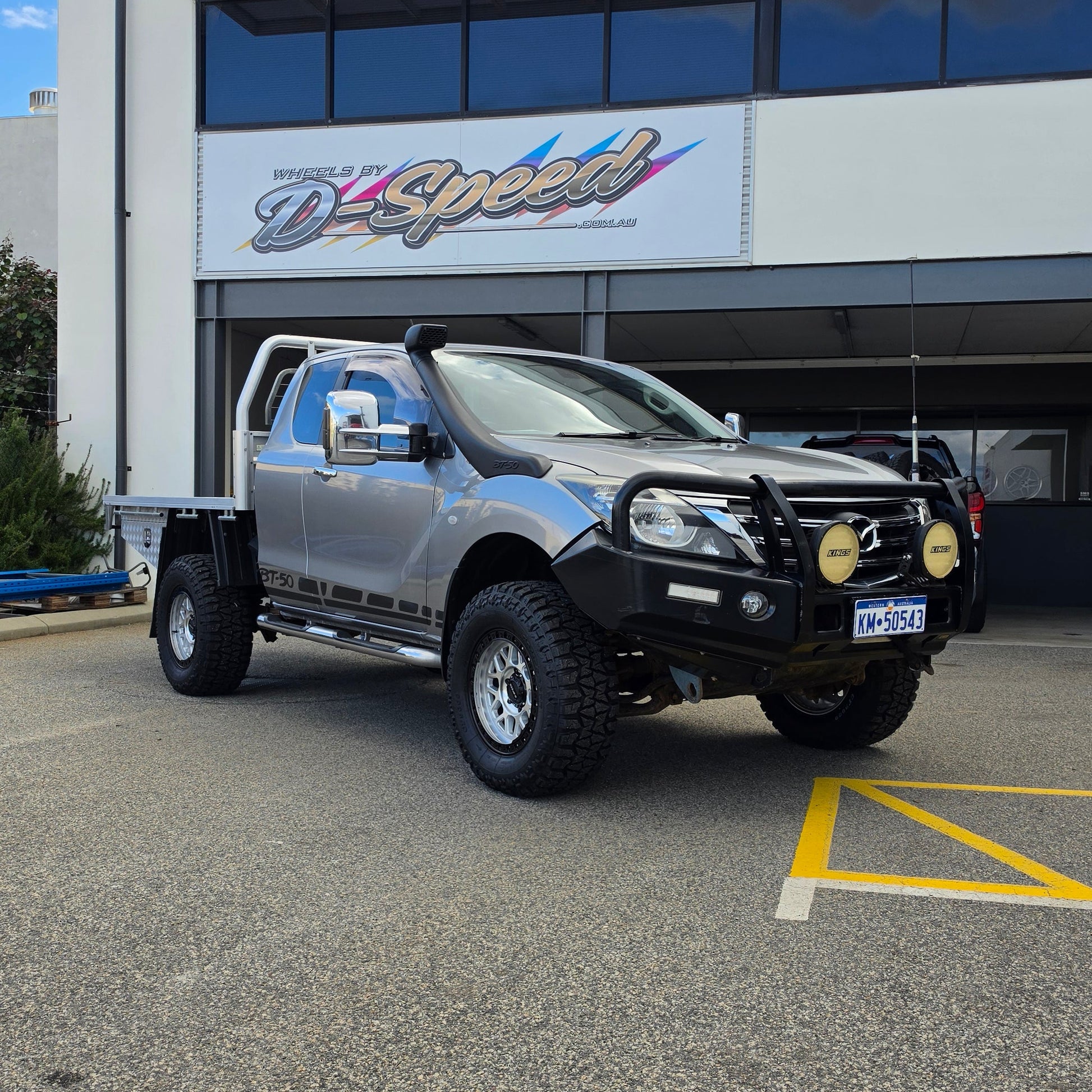 A dark gray off-road BT-50 with DSpeed MH-1 D-Speed MH1 4x4 off road offroad wheels 17x8.5 machined silver black Ring custom flatbed and front bumper guard parked in a paved lot in front of a beige commercial building under a blue sky.