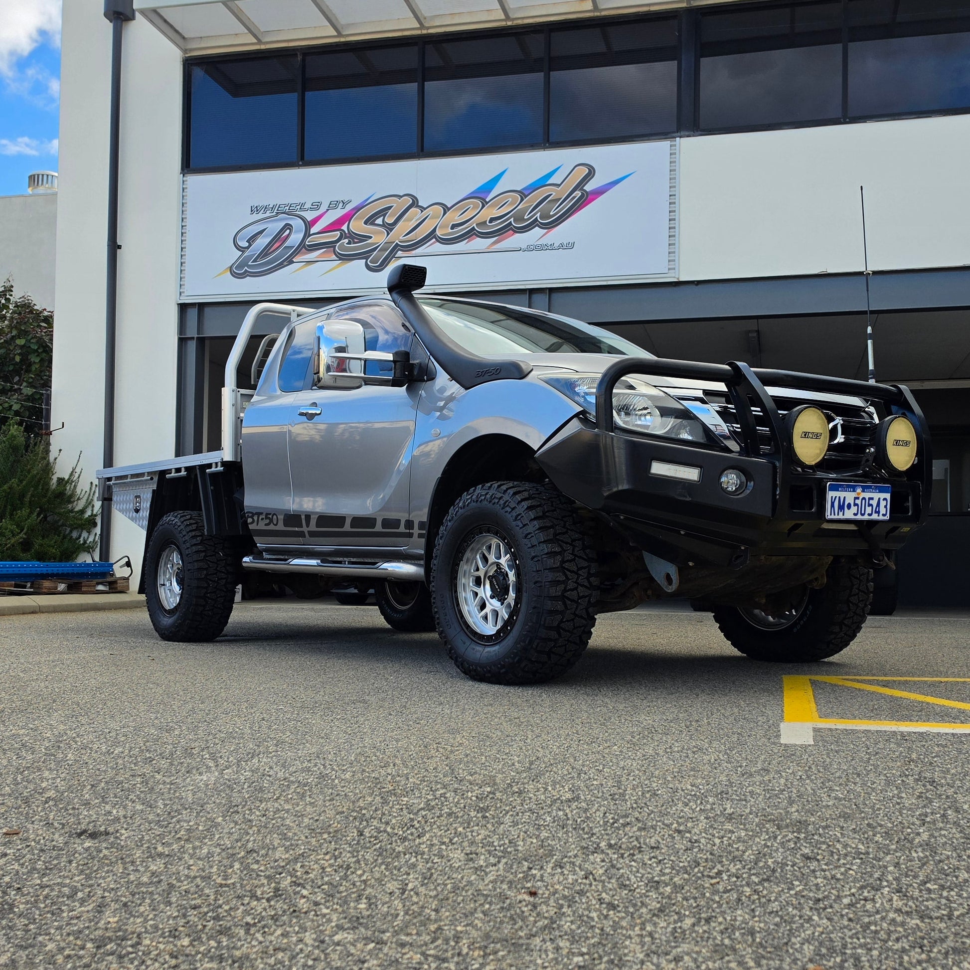 A silver lifted pickup truck with a black bull bar and off-road tires parked in front of a building with a d-speed sign. dspeed mh1 mh-1 alloy wheels 4x4 rims and comforser tyres 17x8.5 machined silver black ring