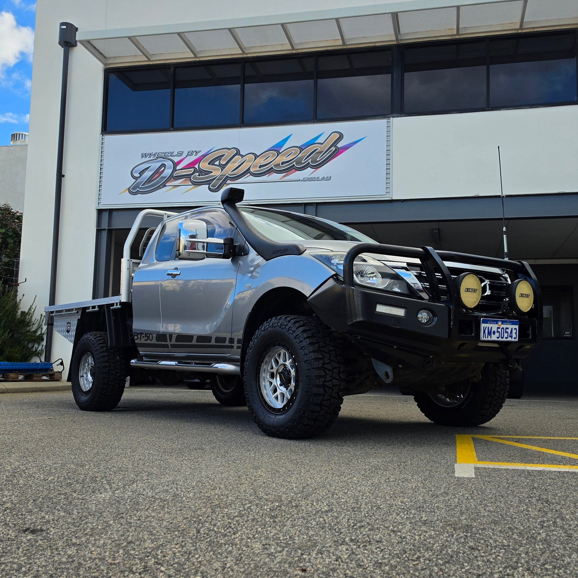 A dark gray off-road BT-50 with DSpeed MH-1 D-Speed MH1 4x4 off road offroad wheels 17x8.5 machined silver black Ring custom flatbed and front bumper guard parked in a paved lot in front of a beige commercial building under a blue sky.