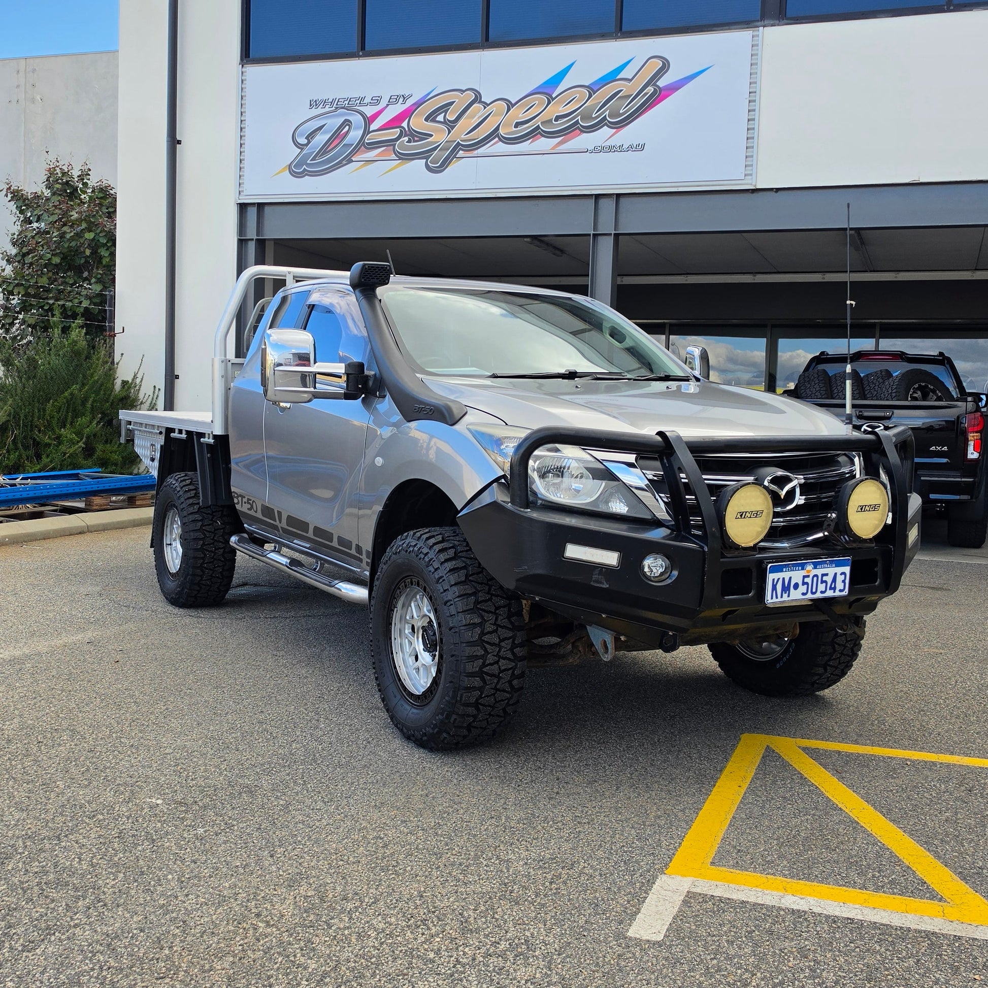 A silver lifted pickup truck with a black bull bar and off-road tires parked in front of a building with a d-speed sign. dspeed mh1 mh-1 alloy wheels 4x4 rims and comforser tyres 17x8.5 machined silver black ring