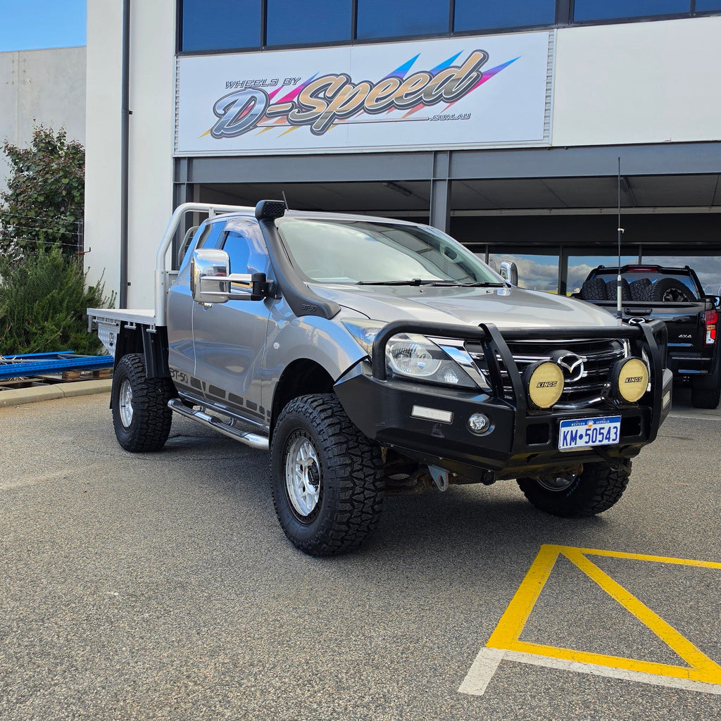 A dark gray off-road BT-50 with DSpeed MH-1 D-Speed MH1 4x4 off road offroad wheels 17x8.5 machined silver black Ring custom flatbed and front bumper guard parked in a paved lot in front of a beige commercial building under a blue sky.