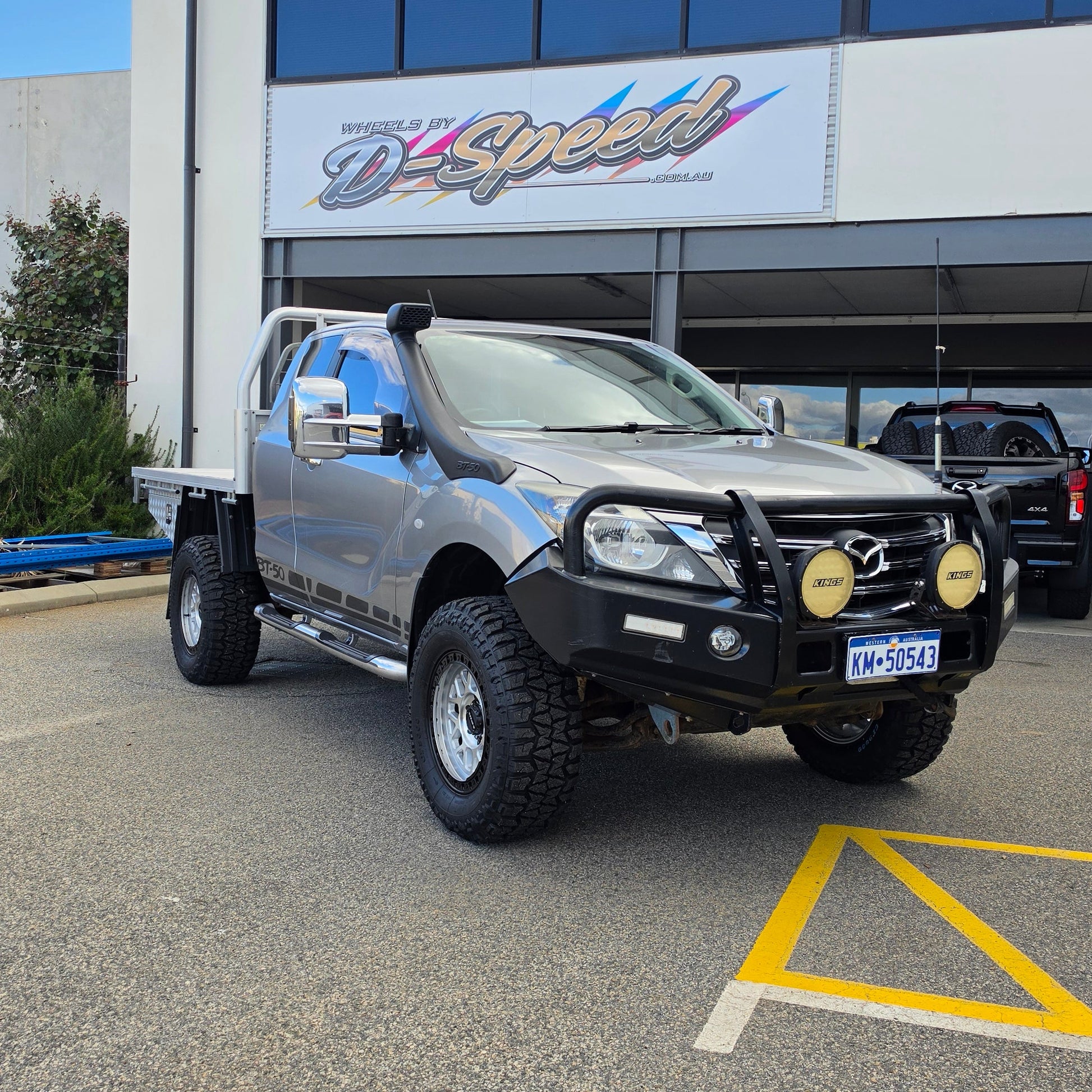 A dark gray off-road BT-50 with DSpeed MH-1 D-Speed MH1 4x4 off road offroad wheels 17x8.5 machined silver black Ring custom flatbed and front bumper guard parked in a paved lot in front of a beige commercial building under a blue sky.