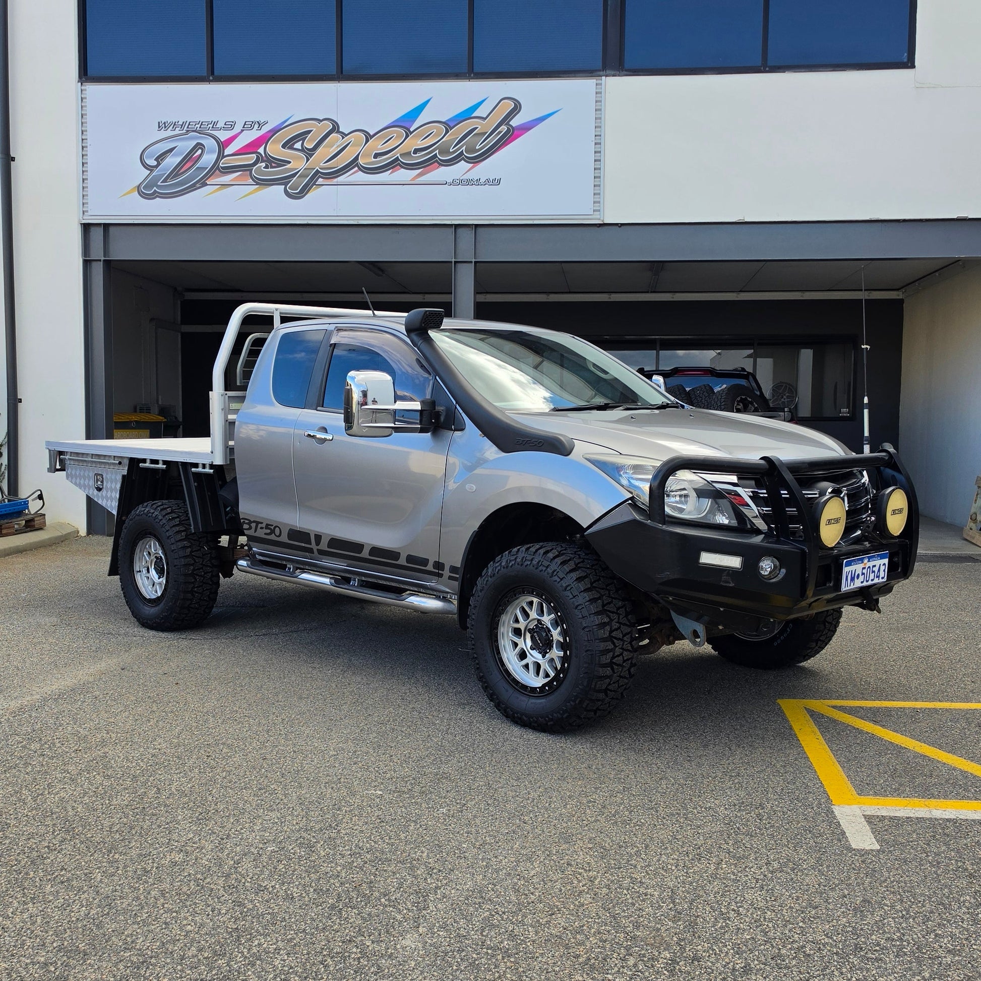 A silver lifted pickup truck with a black bull bar and off-road tires parked in front of a building with a d-speed sign. dspeed mh1 mh-1 alloy wheels 4x4 rims and comforser tyres 17x8.5 machined silver black ring