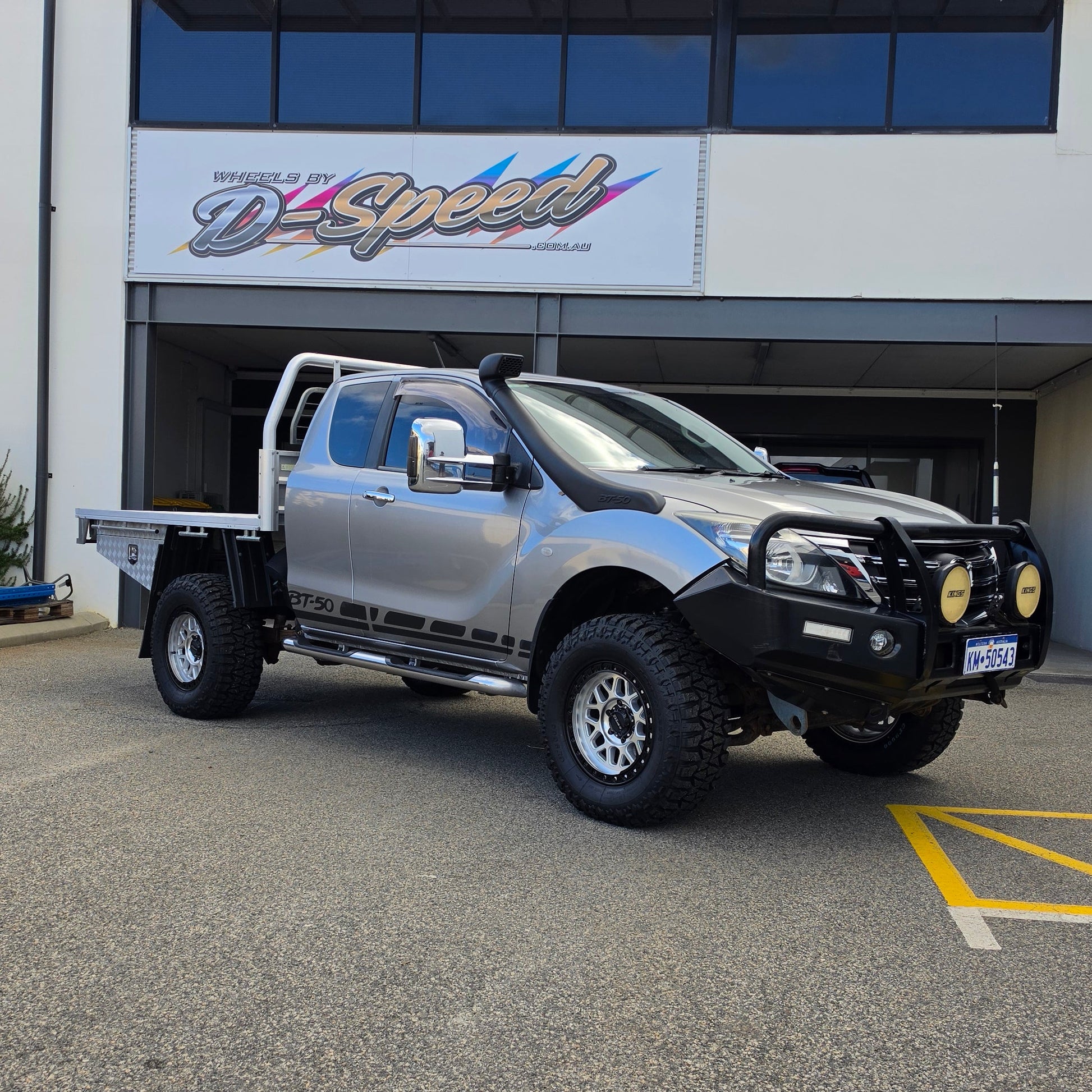 A dark gray off-road BT-50 with DSpeed MH-1 D-Speed MH1 4x4 off road offroad wheels 17x8.5 machined silver black Ring custom flatbed and front bumper guard parked in a paved lot in front of a beige commercial building under a blue sky.