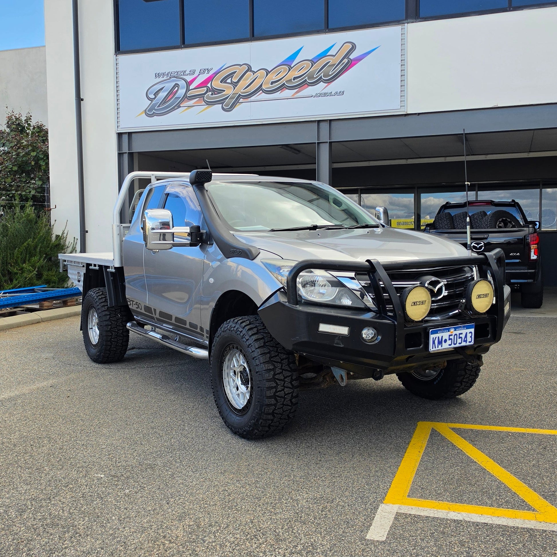 A dark gray off-road BT-50 with DSpeed MH-1 D-Speed MH1 4x4 off road offroad wheels 17x8.5 machined silver black Ring custom flatbed and front bumper guard parked in a paved lot in front of a beige commercial building under a blue sky.