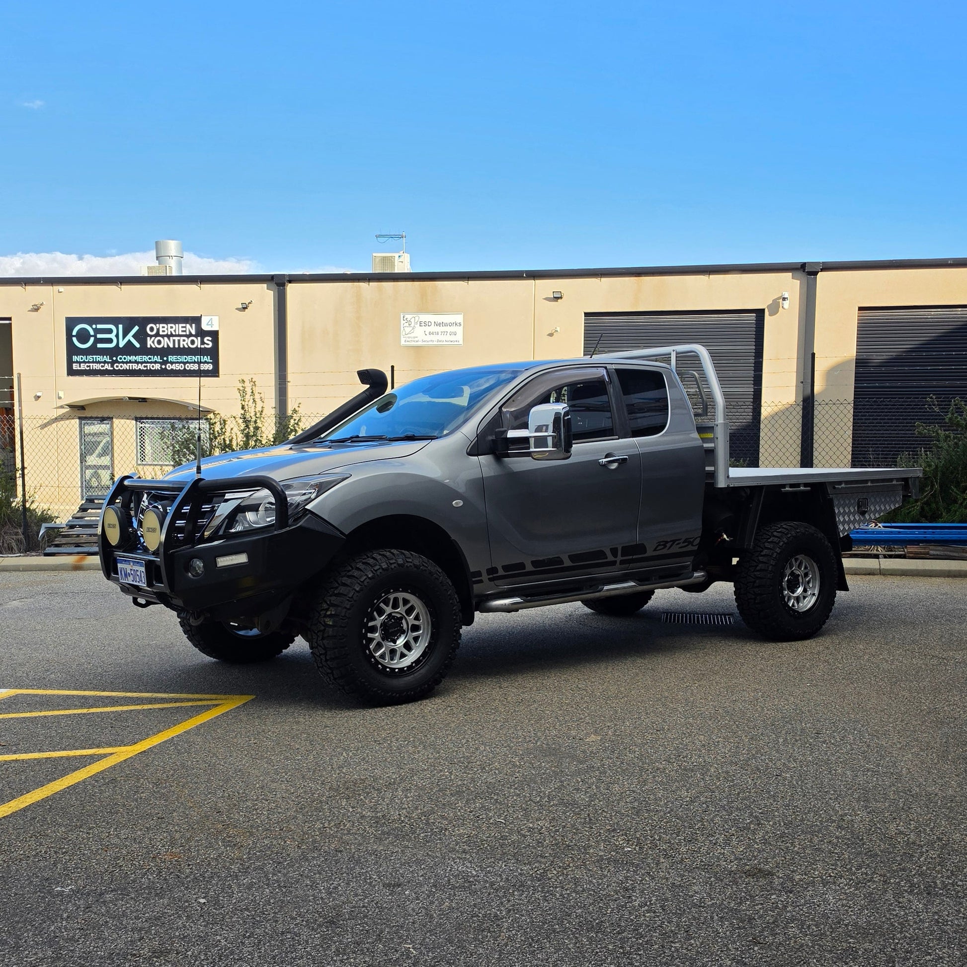 A dark gray off-road BT-50 with DSpeed MH-1 D-Speed MH1 4x4 off road offroad wheels 17x8.5 machined silver black Ring custom flatbed and front bumper guard parked in a paved lot in front of a beige commercial building under a blue sky.