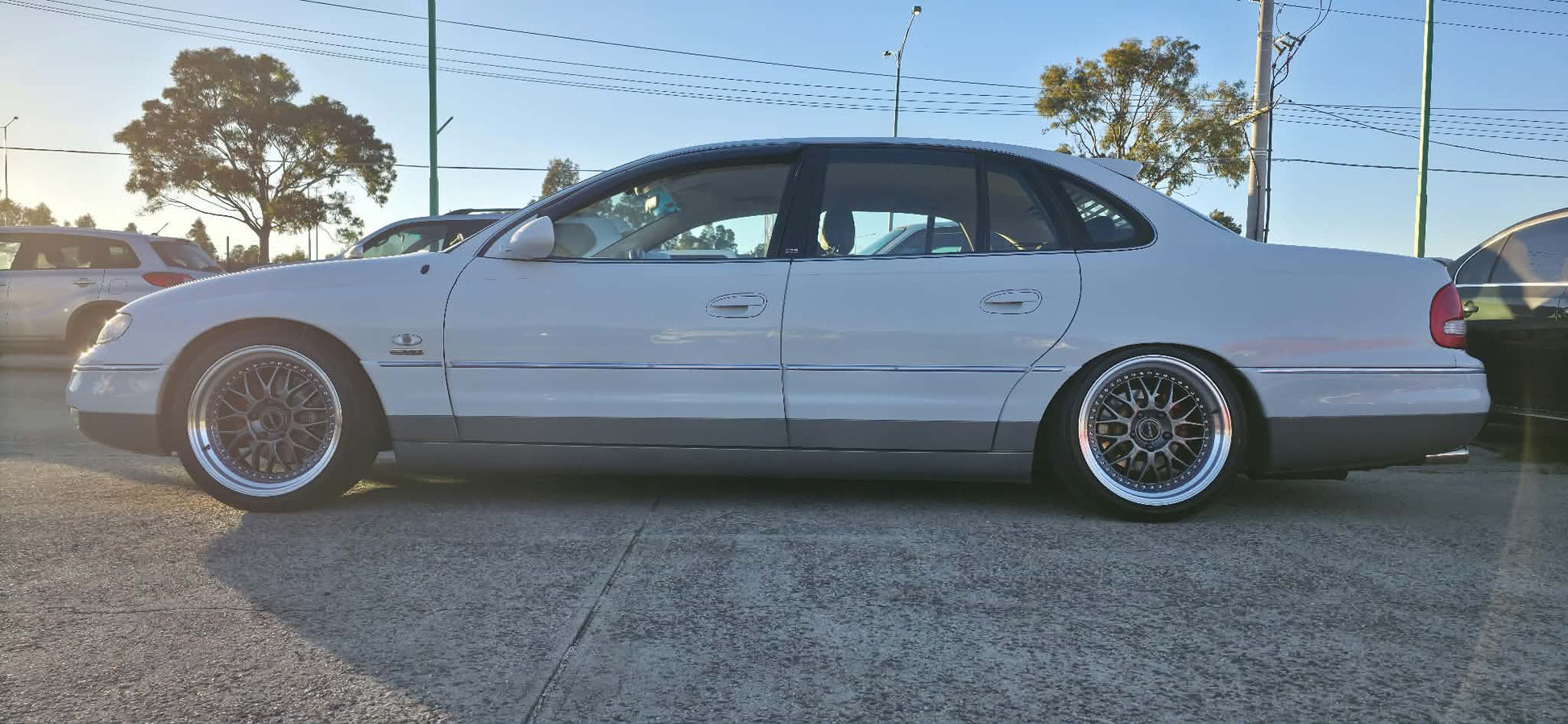 A white holden Statesman parked outdoors on a gravel lot, featuring a lowered suspension, custom multi-spoke alloy wheels, and a tinted rear window. Dspeed d-speed ds06 ds-06 19inch 19x8.5 19x9.5 wheels with tyres dish wheels dished rims