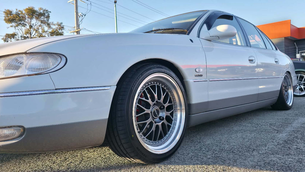 A white holden Statesman parked outdoors on a gravel lot, featuring a lowered suspension, custom multi-spoke alloy wheels, and a tinted rear window. Dspeed d-speed ds06 ds-06 19inch 19x8.5 19x9.5 wheels with tyres dish wheels dished rims