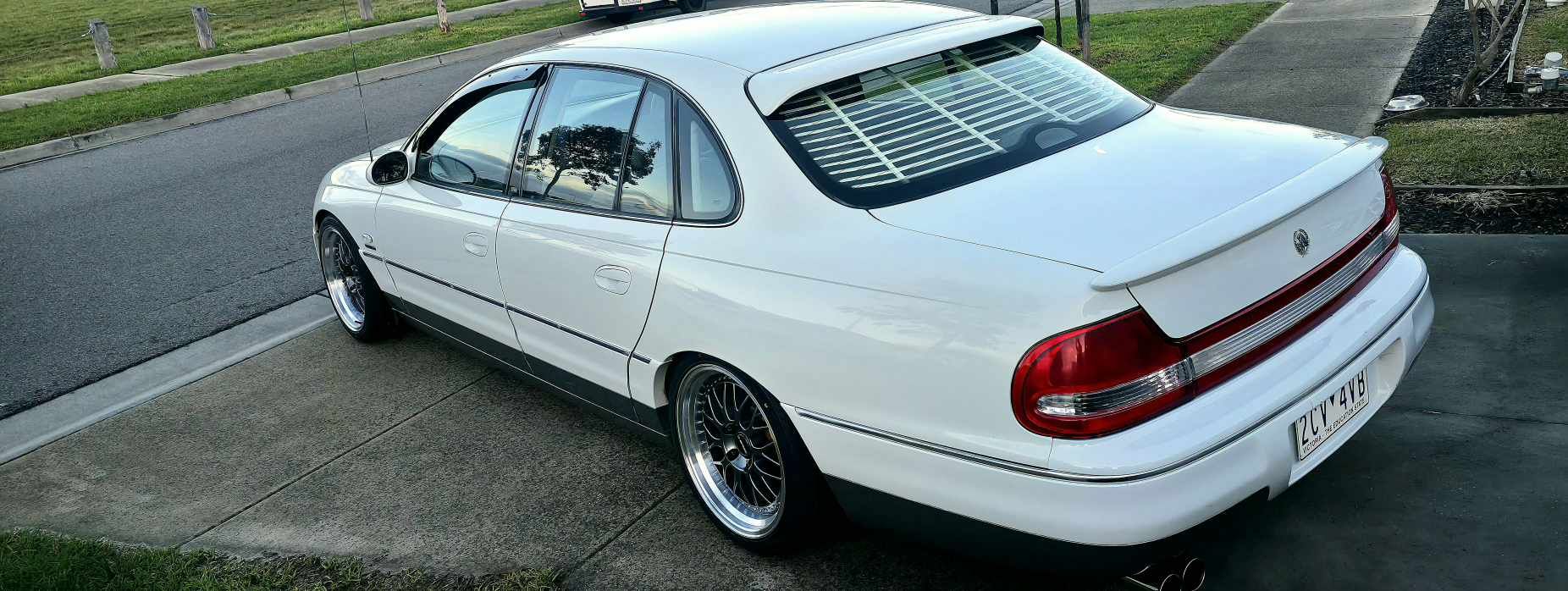 A white holden Statesman parked outdoors on a gravel lot, featuring a lowered suspension, custom multi-spoke alloy wheels, and a tinted rear window. Dspeed d-speed ds06 ds-06 19inch 19x8.5 19x9.5 wheels with tyres dish wheels dished rims