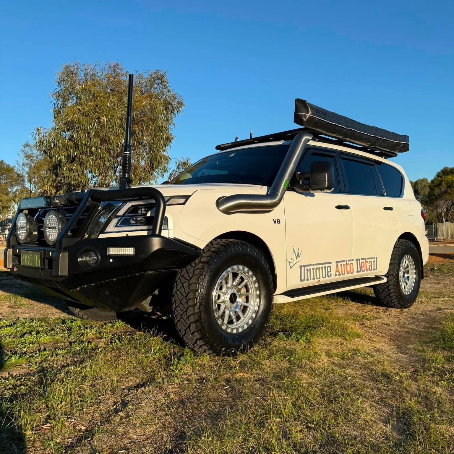 A white nissan patrol Y62 off-road SUV parked outdoors on grassy ground under a blue sky, with a black roof rack, front bull bar, and off-road tires. DSpeed d-speed XT-1 xt1 18x9 wheels 4x4 rims off road offroad