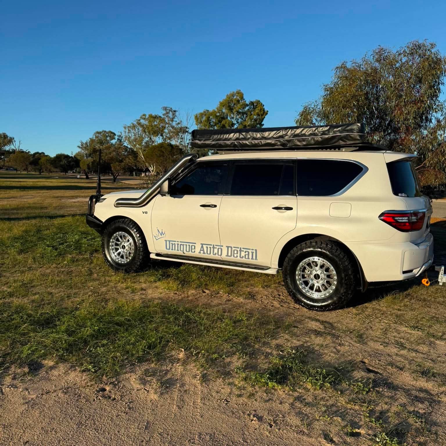 A white nissan patrol Y62 off-road SUV parked outdoors on grassy ground under a blue sky, with a black roof rack, front bull bar, and off-road tires. DSpeed d-speed XT-1 xt1 18x9 wheels 4x4 rims off road offroad