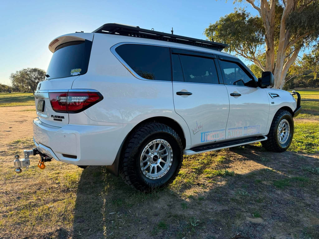 A white nissan patrol Y62 off-road SUV parked outdoors on grassy ground under a blue sky, with a black roof rack, front bull bar, and off-road tires. DSpeed d-speed XT-1 xt1 18x9 wheels 4x4 rims off road offroad