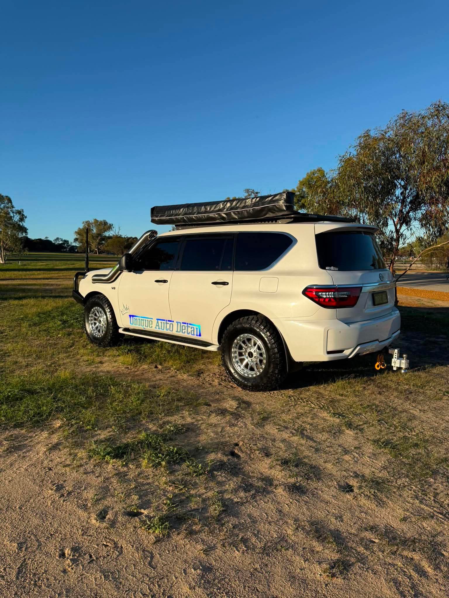 A white nissan patrol Y62 off-road SUV parked outdoors on grassy ground under a blue sky, with a black roof rack, front bull bar, and off-road tires. DSpeed d-speed XT-1 xt1 18x9 wheels 4x4 rims off road offroad