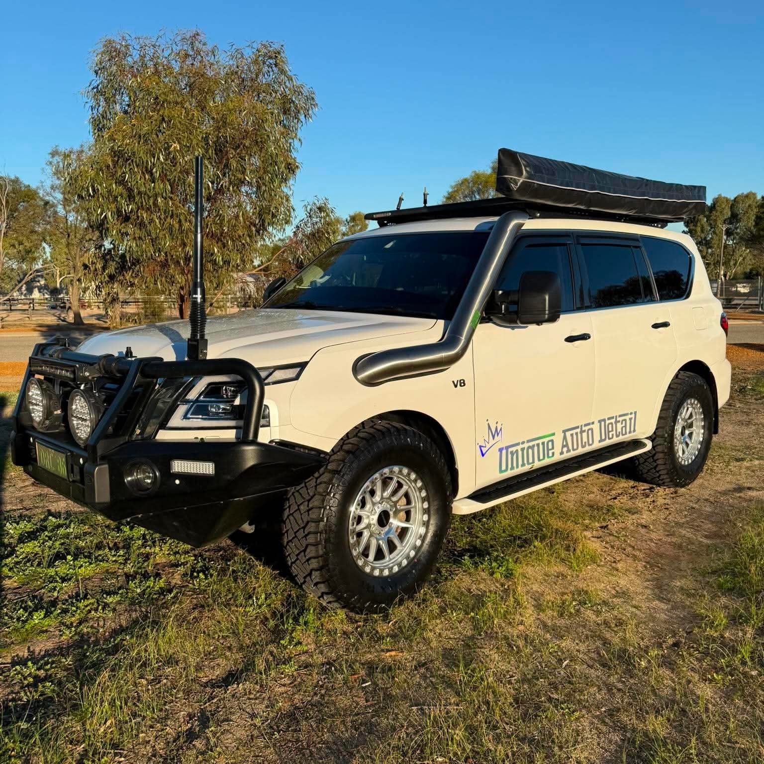 A white nissan patrol Y62 off-road SUV parked outdoors on grassy ground under a blue sky, with a black roof rack, front bull bar, and off-road tires. DSpeed d-speed XT-1 xt1 18x9 wheels 4x4 rims off road offroad